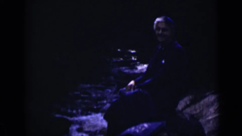 1938 Colorado frame showing an adult seated at the edge of a rocky stream under dark blue-tinted exposure.