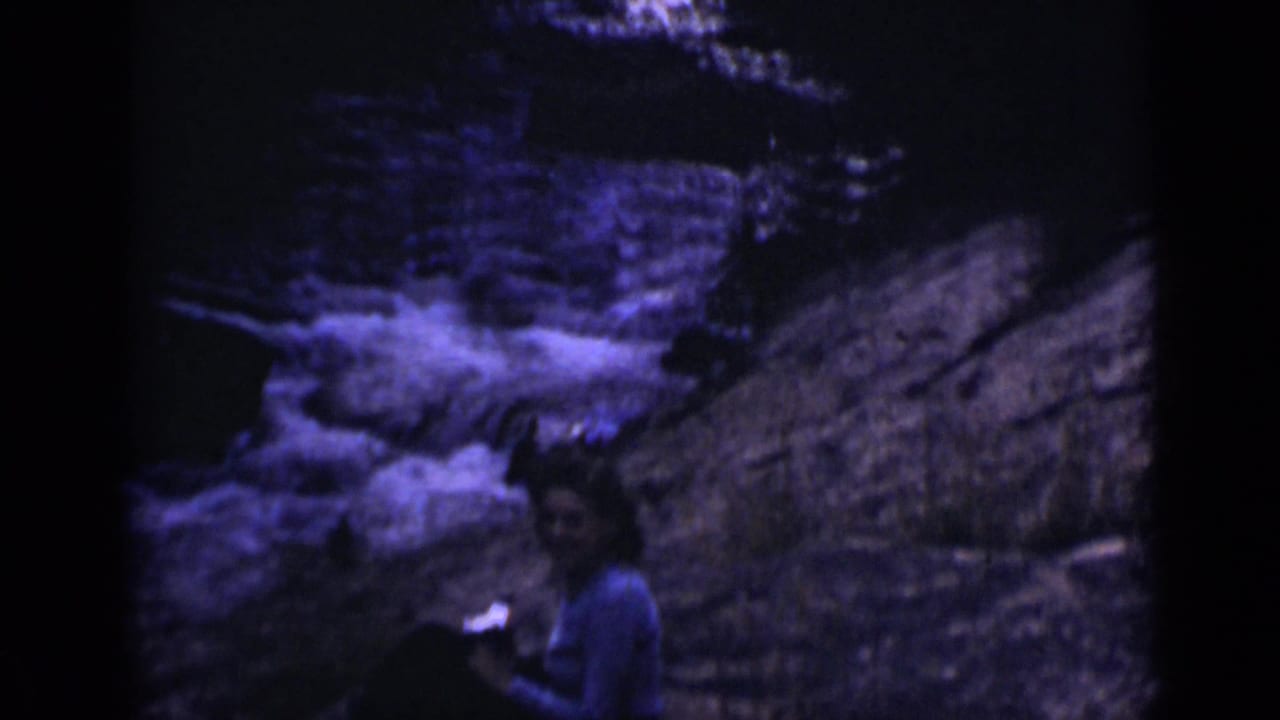A seated person remains on the rocks near rushing water in Colorado archival 8mm footage.
