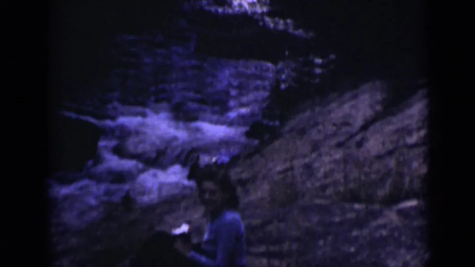 A seated person remains on the rocks near rushing water in Colorado archival 8mm footage.