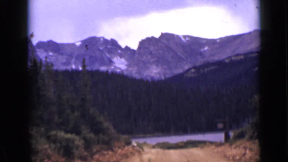 Wide 1938 Colorado landscape view of a lake, evergreen forest, and distant snow-dusted mountains.
