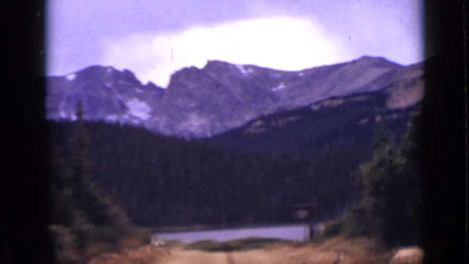 Archival Colorado 8mm frame showing a lake below forested slopes and a rugged mountain ridge.