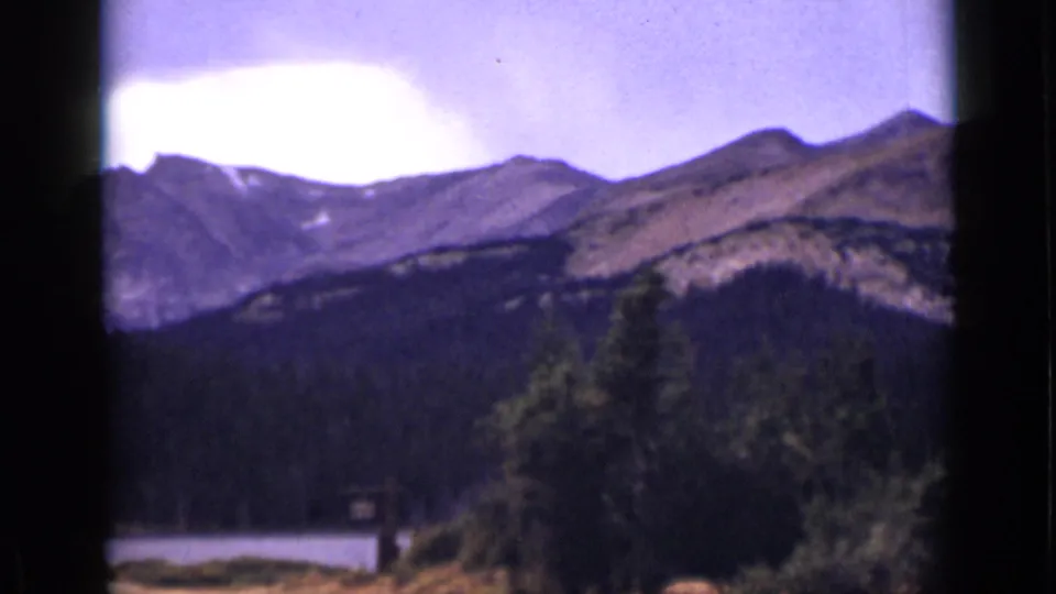 Colorado mountain vista on 8mm film, with dark forest, pale sky, and a lake partly visible near the lower left.