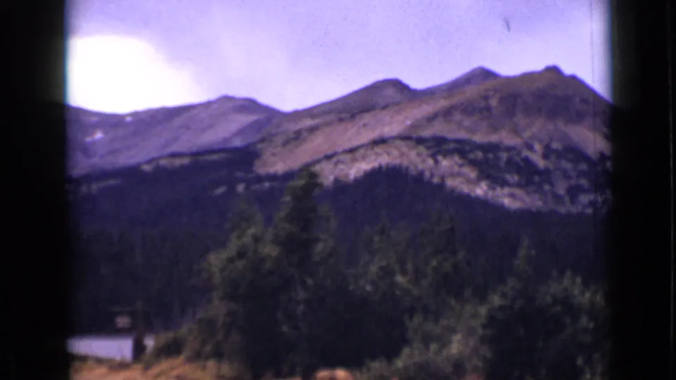 1938 Colorado scenic frame showing a rocky ridge, evergreen forest, foreground brush, and a small visible section of lake.