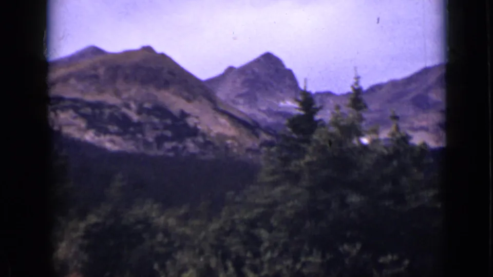 Colorado mountain landscape on 8mm film, with evergreen trees in the foreground and hazy peaks beneath a pale sky.