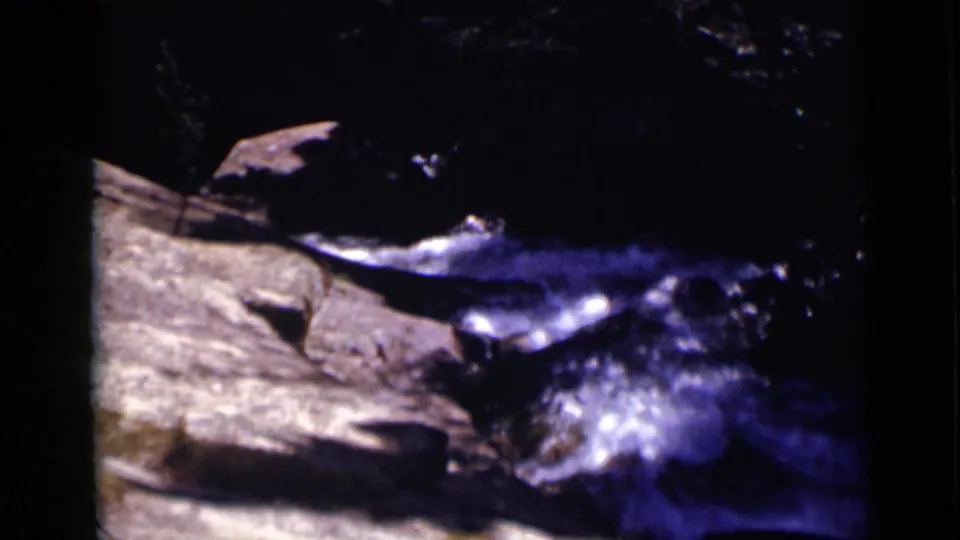 Vintage 8mm view of a shaded Colorado creek where bright water moves along a broad rock surface.