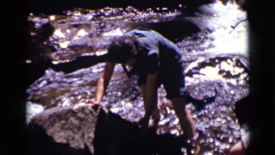 A person in dark shirt and shorts appears to steady themself on a large rock while crossing a Colorado stream in 1938.