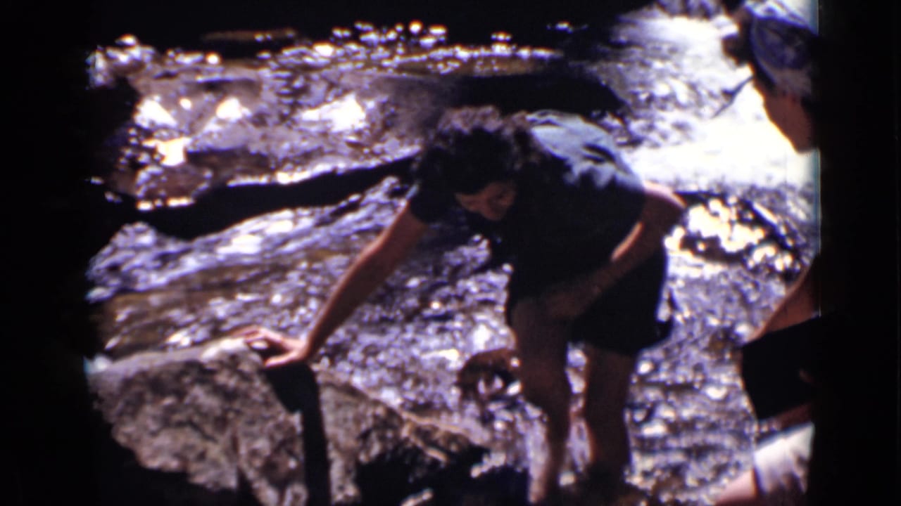 Woman crouching beside a rocky stream with one hand near the water.