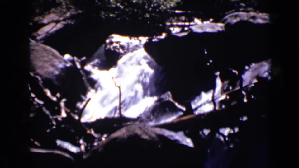 A rocky stream cutaway shows bright moving water, shaded stones, and branch shapes in 1938 Colorado home-movie footage.