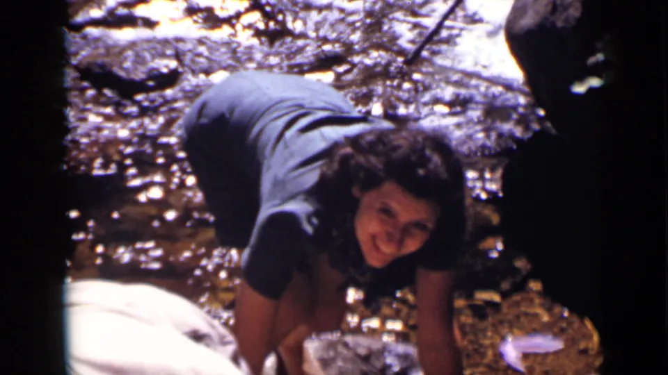 A woman crouches by the rocky stream and smiles toward the camera in the supported Colorado 1938 home-movie scene.