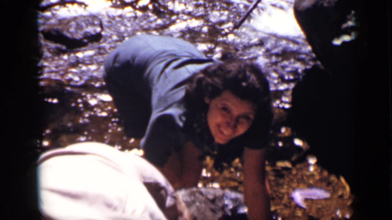 In 1938 Colorado home-movie footage, a woman crouches near a sunlit stream as seated companions rest in the wooded background.