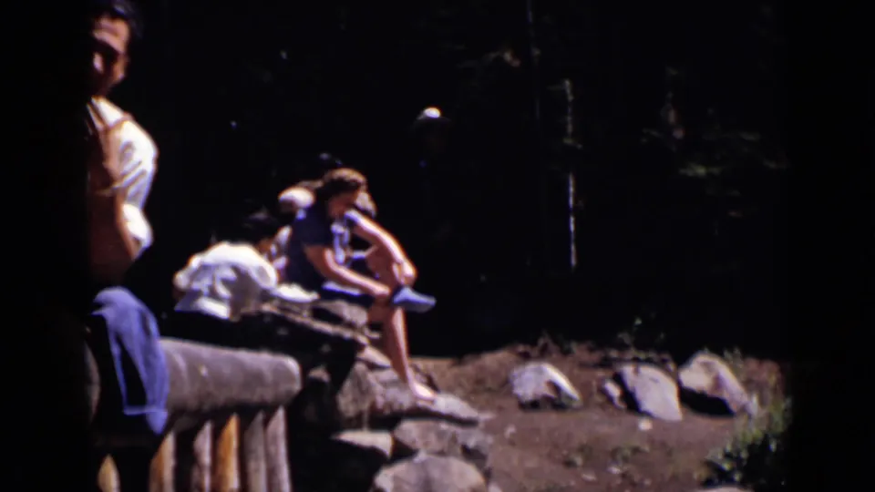 A small group rests on a log bench in a wooded Colorado setting, with rocks and deep shade around the clearing.