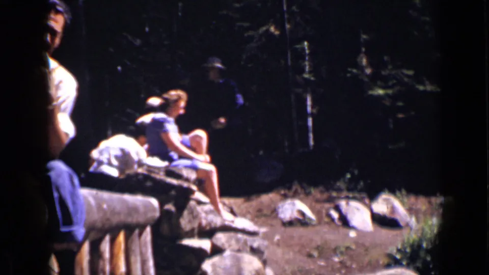 The home movie frames companions seated on a rustic bench in a shaded Colorado forest area, with rocks scattered in front.
