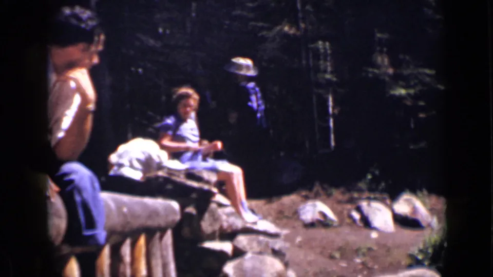 In the wooded Colorado scene, a woman sits on a log bench with companions partly visible at the edge of the 8mm frame.