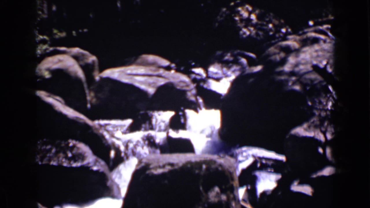 Water rushes between dark rocks in a shaded forest setting.