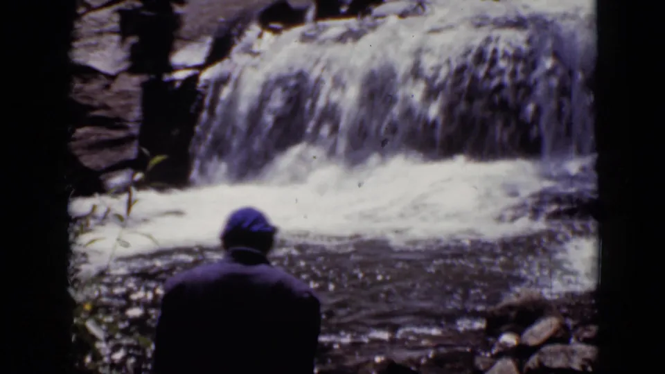 1938 Colorado home-movie frame with a woman in dark outerwear and blue head covering standing before a low waterfall.