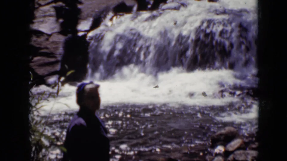 1938 Colorado frame showing a woman in profile near the stream edge as water falls over rocks in the background.