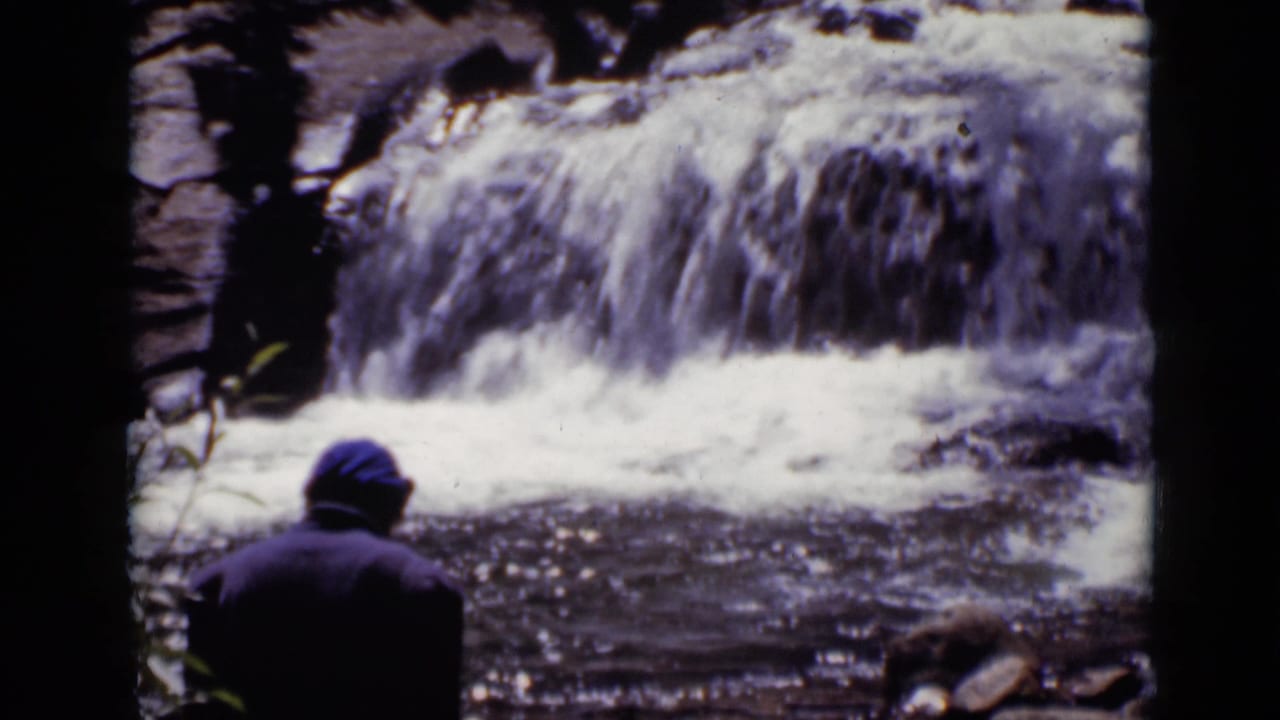 In 1938 Colorado home-movie footage, a person in a blue cap is seen from behind near a rushing rocky waterfall.