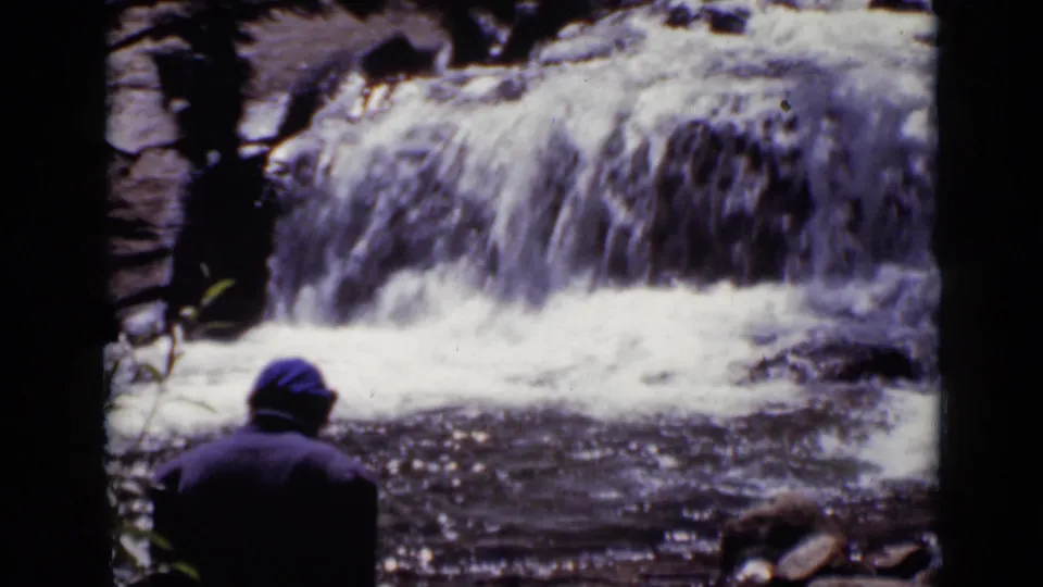 In 1938 Colorado home-movie footage, a person in a blue cap is seen from behind near a rushing rocky waterfall.