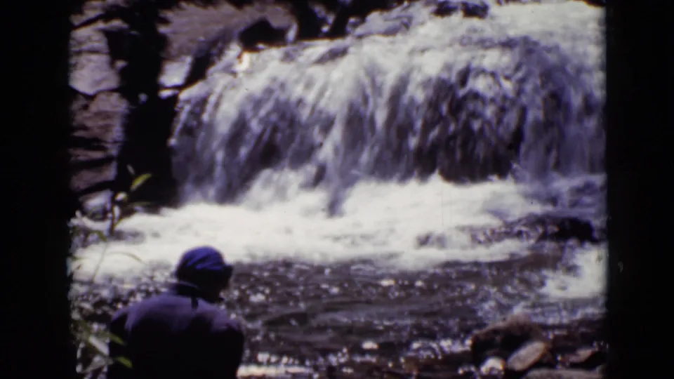 A person in a blue cap and dark jacket looks toward a bright waterfall in Colorado, with rocks and stream water in view.