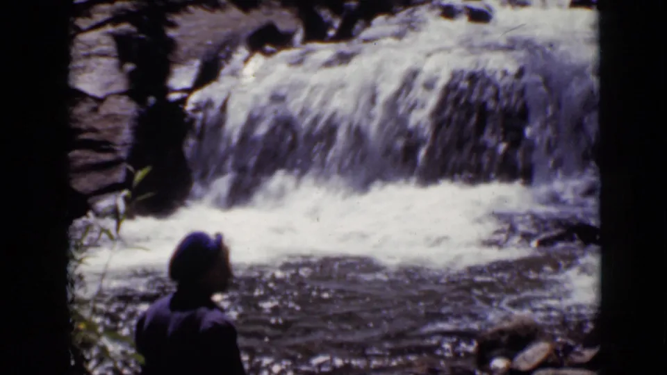 The visitor appears in partial profile beside the rushing waterfall, framed by rocks and foliage in 1938 Colorado footage.