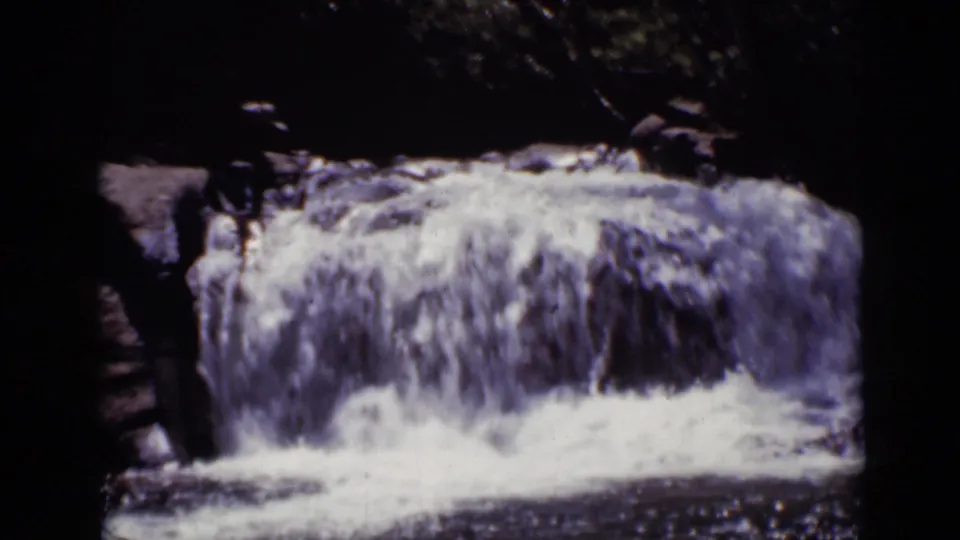1938 Colorado 8mm frame showing a small waterfall dropping over rocks into foamy water, with dark trees and foliage around the stream.