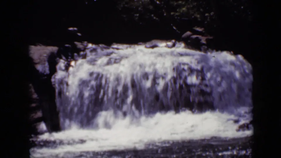 1938 Colorado 8mm frame with bright falling water and foam beneath a small rock ledge, bordered by dark forest shadows.