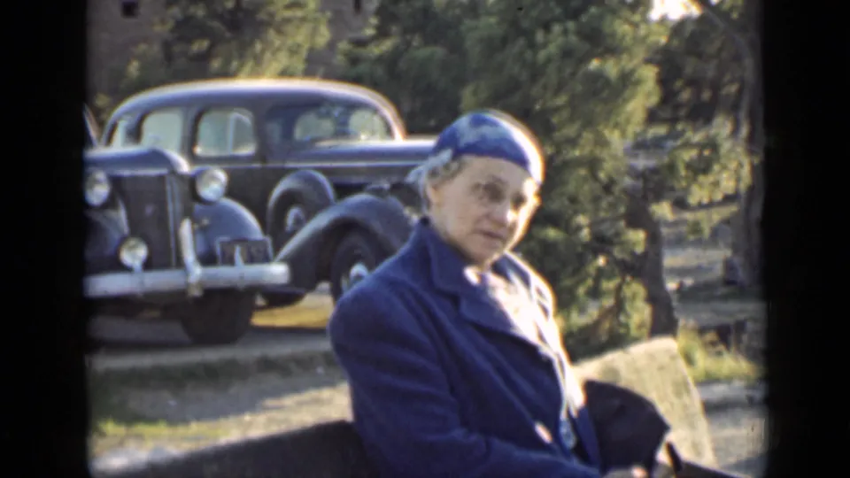 In Arizona home-movie footage from 1939, a woman in a blue coat and hat stands near two parked black cars and trees.