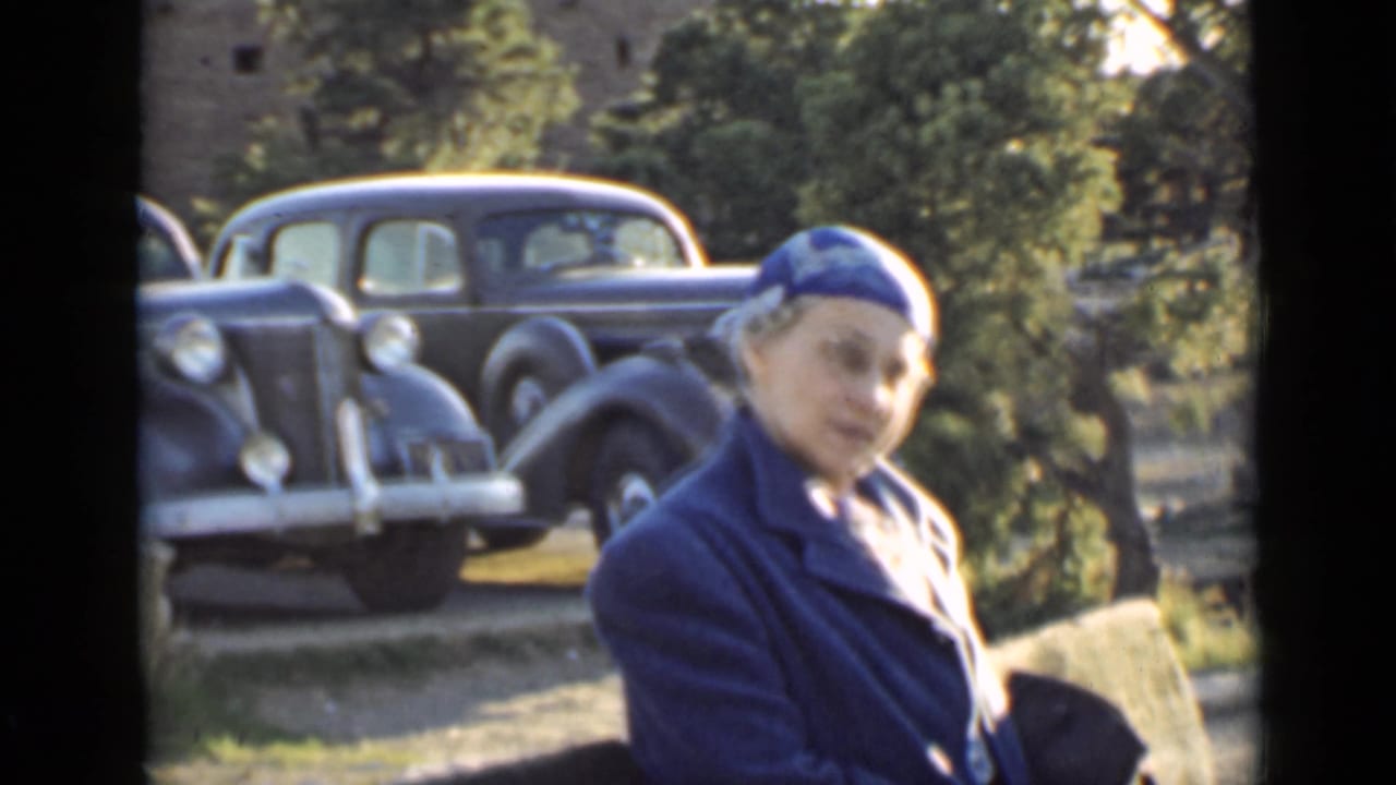1939 Arizona home-movie frame showing a woman in a blue coat and hat near 1930s automobiles and a stone structure.