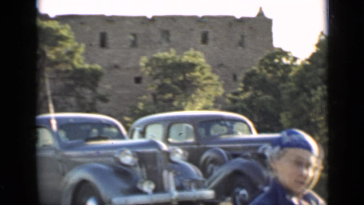 Classic automobile parked near a stone building with a woman partly visible at right.