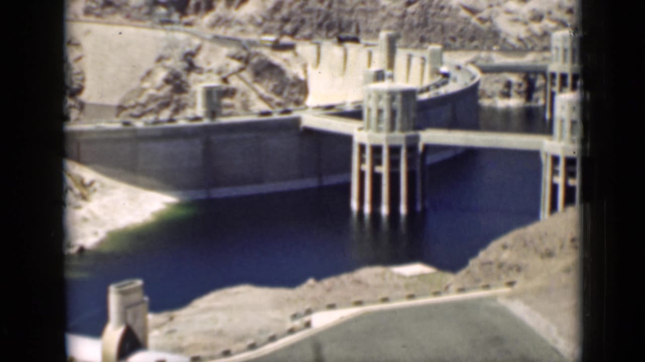 Concrete dam and intake towers beside dark reservoir water in rocky desert terrain.