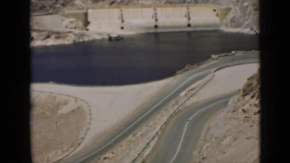 1939 Arizona 8mm frame looking across a reservoir edge toward a concrete dam wall and a winding road in desert terrain.