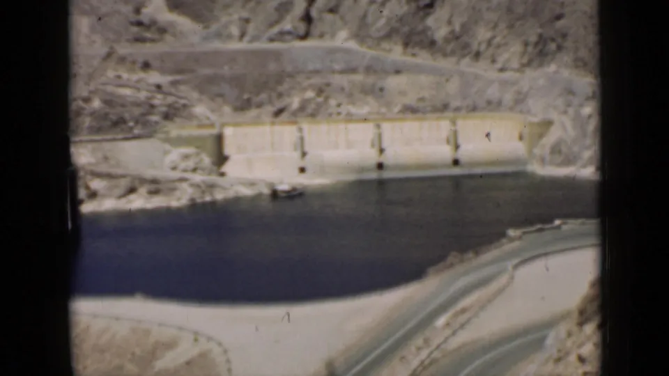 1939 Arizona 8mm frame showing the dam wall across rocky terrain, with reservoir water and a curving road visible below.