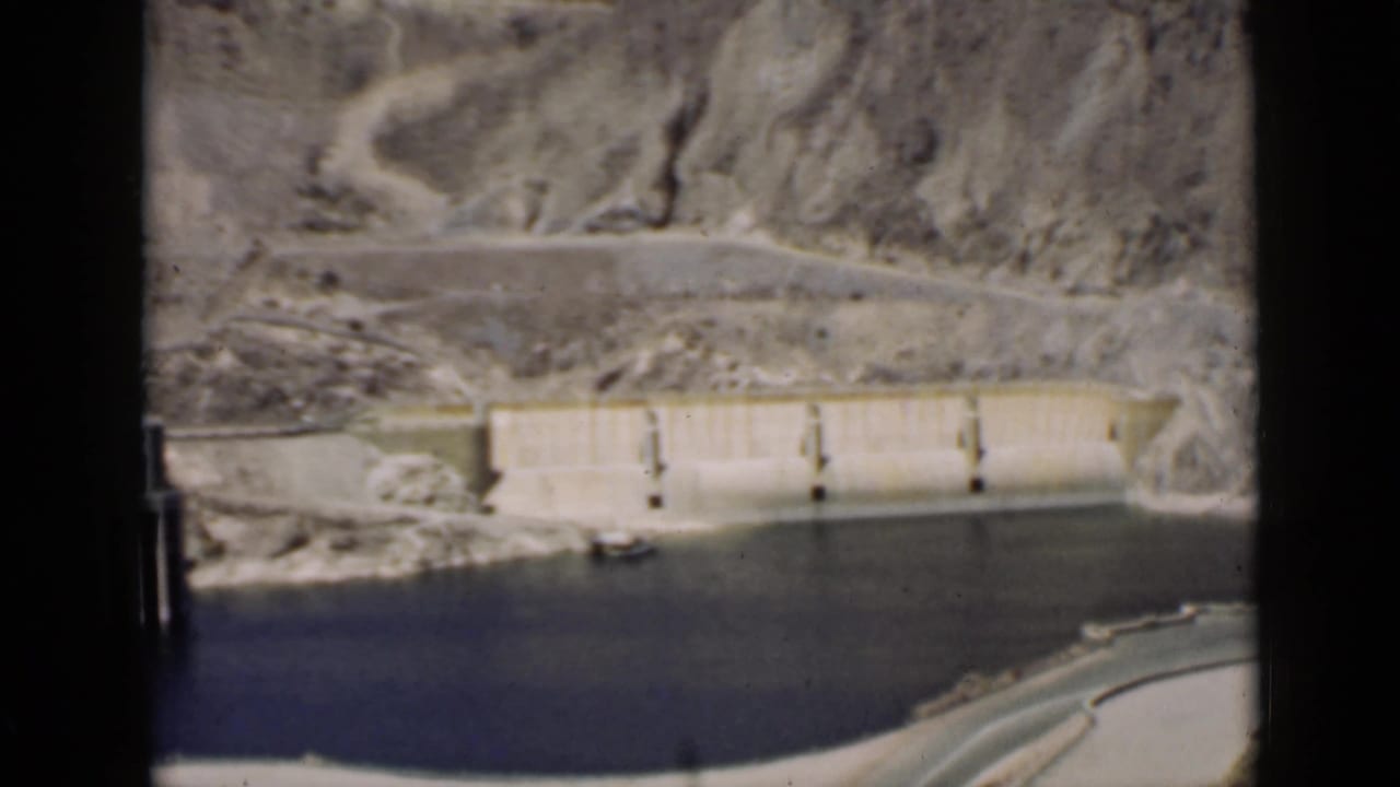 Concrete dam and reservoir below rocky desert hills with a road above.