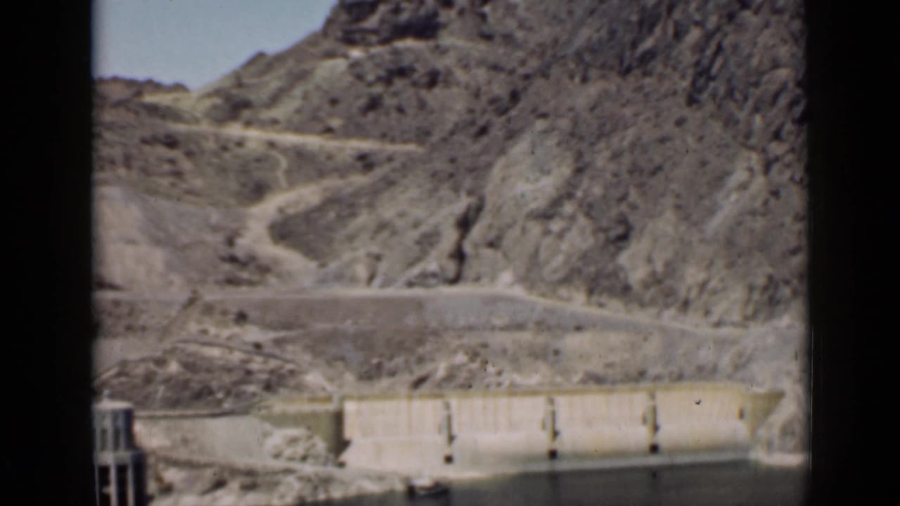 Dam structure beside a reservoir with a winding road across rocky hills.