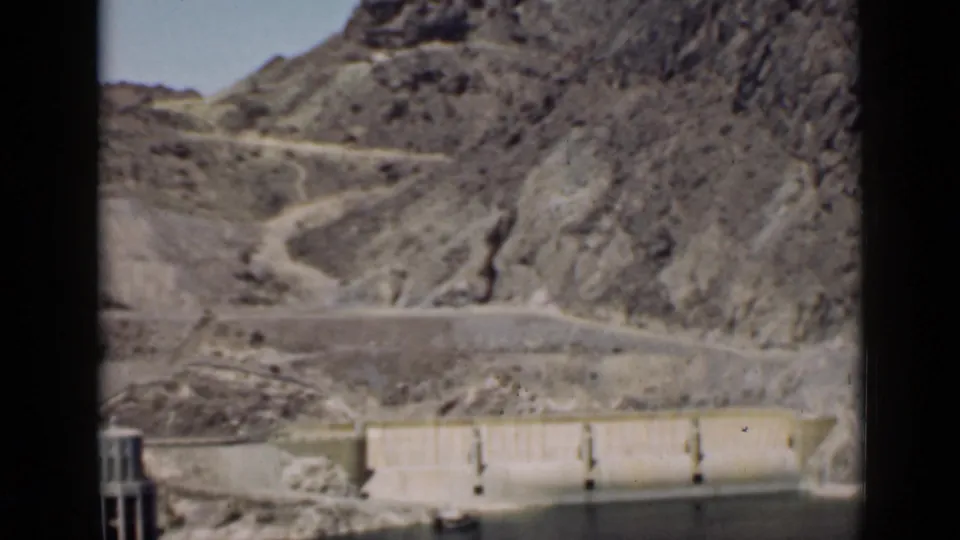 Arizona desert dam scene with reservoir water in the foreground and a winding road climbing through rocky terrain.