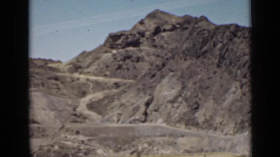 Home-movie frame of rugged Arizona hills and a pale road curving through dry terrain near the dam site.