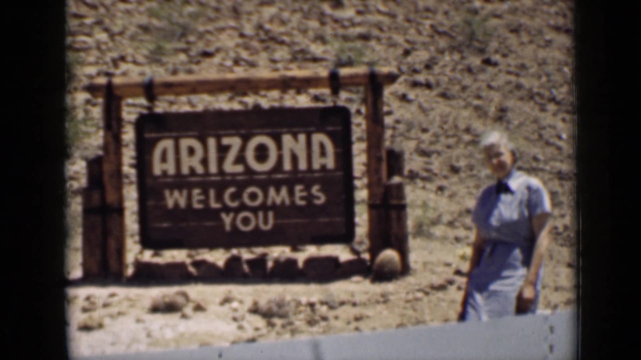 Woman standing beside an Arizona Welcomes You sign in a rocky desert setting.