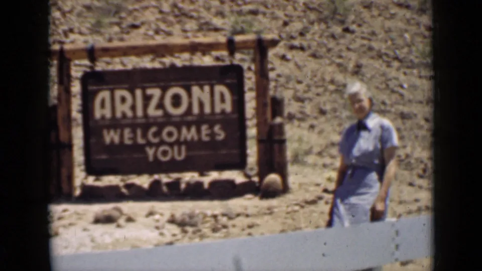 Late-1930s 8mm frame of a woman beside an Arizona Welcomes You sign in a bright arid roadside landscape.