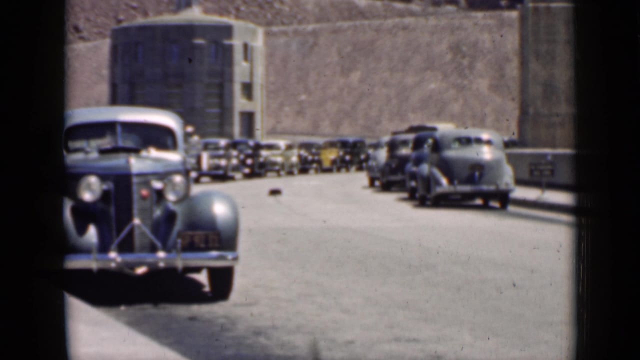 Classic cars lined along a road beside a large concrete structure near rocky terrain.
