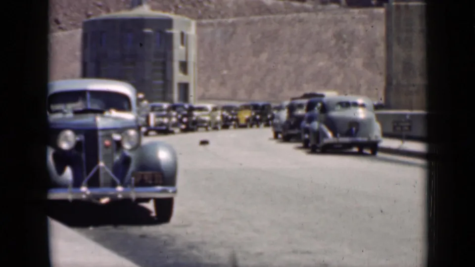 1939 Arizona travel frame with 1930s automobiles parked or moving along a road beside a large concrete dam-like structure.