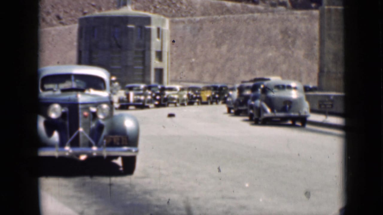 Vintage cars queued on a paved road beside a large concrete structure.