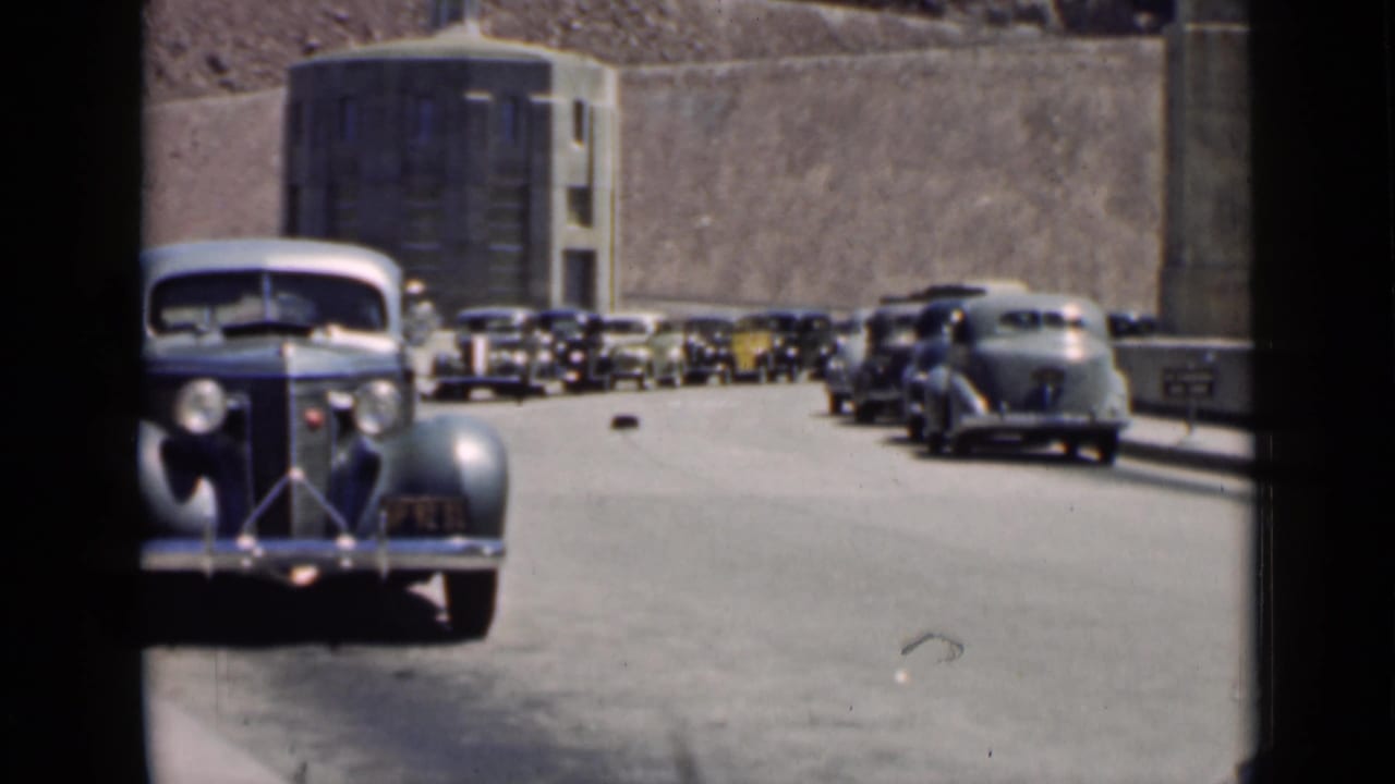 Line of vintage automobiles waiting along a wide road near concrete walls.