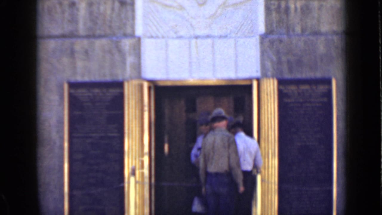 Three men stand at a stone monument entrance with gold-toned doors and dark plaques.