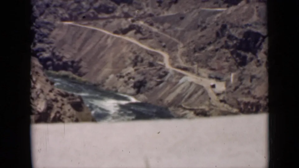 1939 Arizona home-movie frame showing a high view over a rugged canyon, winding road, and river beside a dam structure.