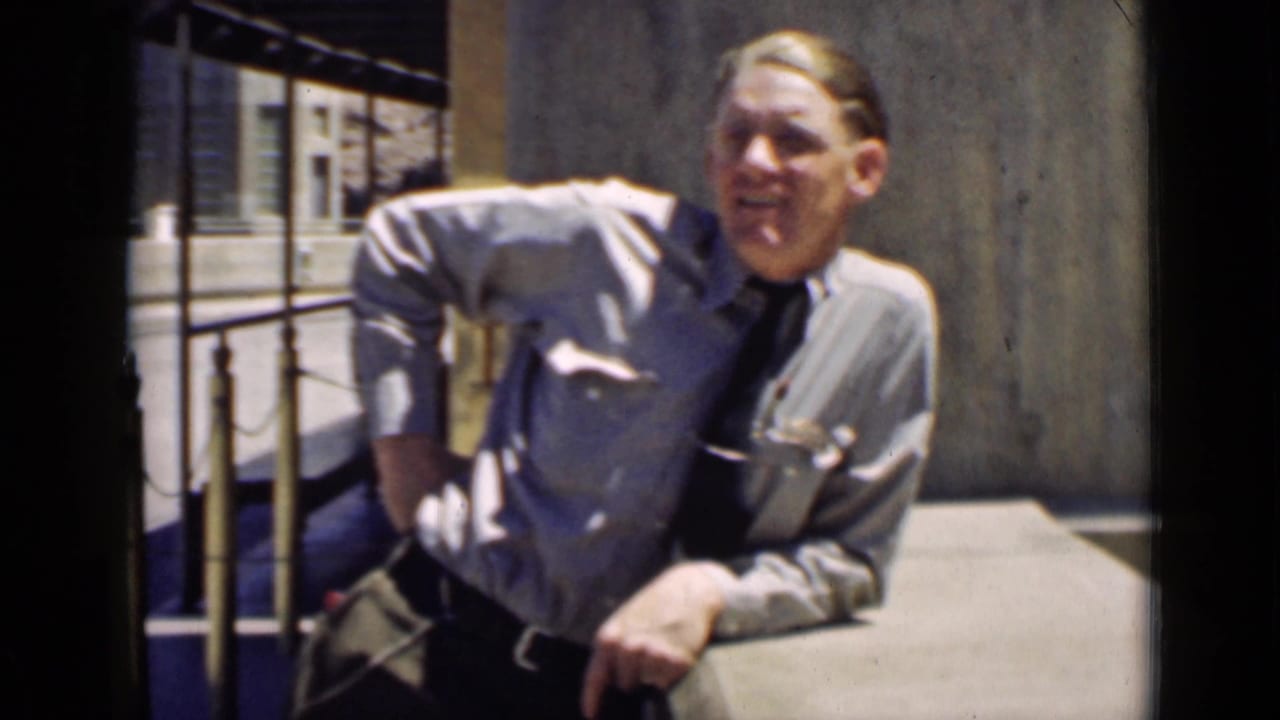 Uniformed blond man leaning on a railing beside a concrete wall.