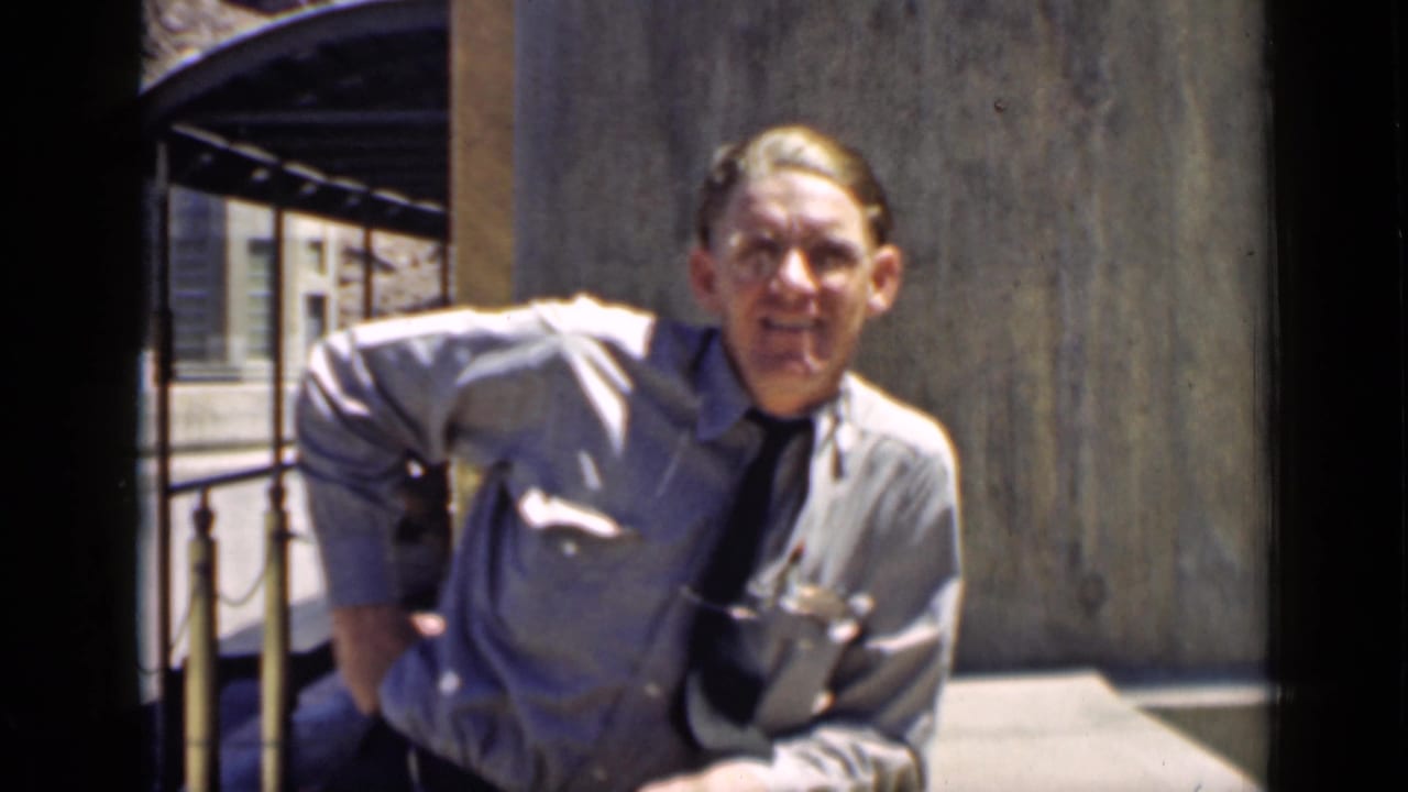 Man in a light shirt and tie smiling near a building exterior.