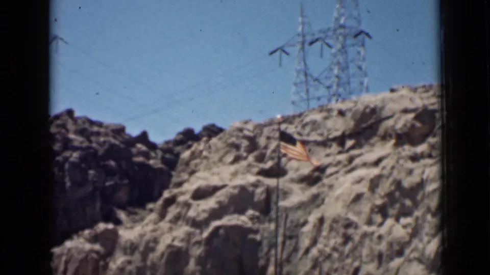 A rocky Arizona slope shows an American flag on a pole, with transmission towers standing on the ridge beyond.