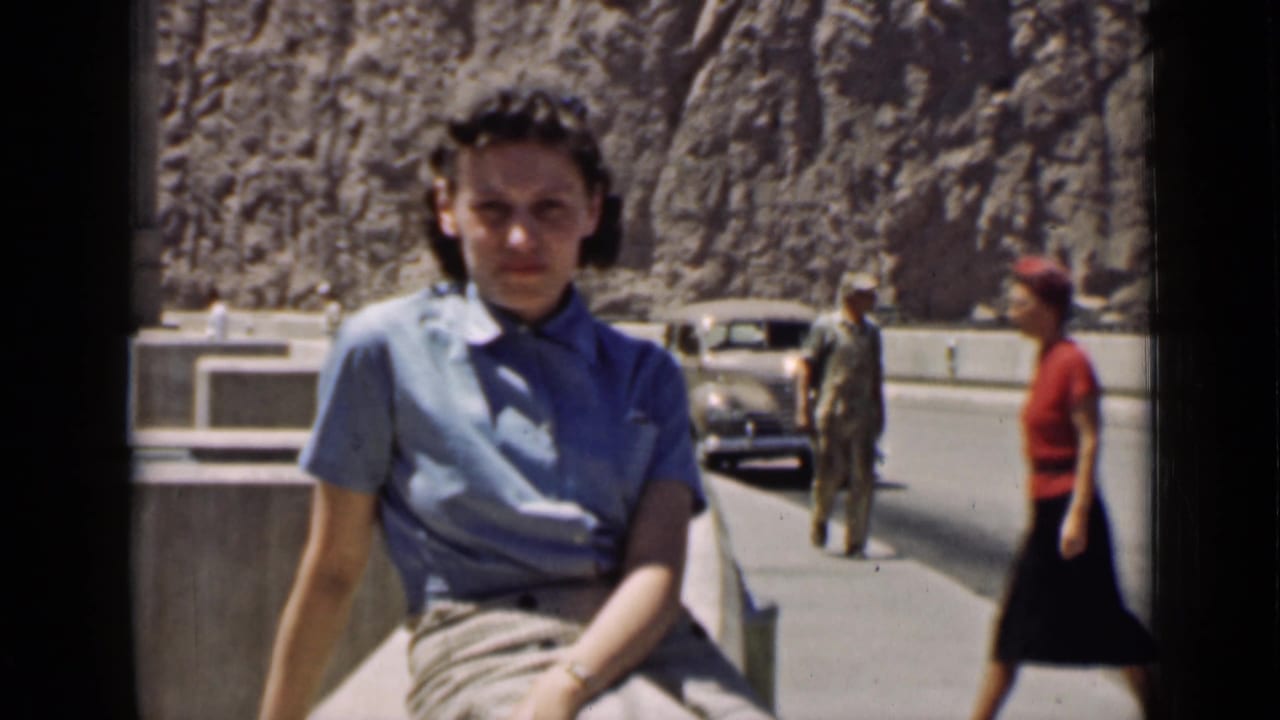 Arizona 1939 frame of a woman in a blue shirt seated on a concrete roadside barrier with pedestrians and a vintage car behind her.