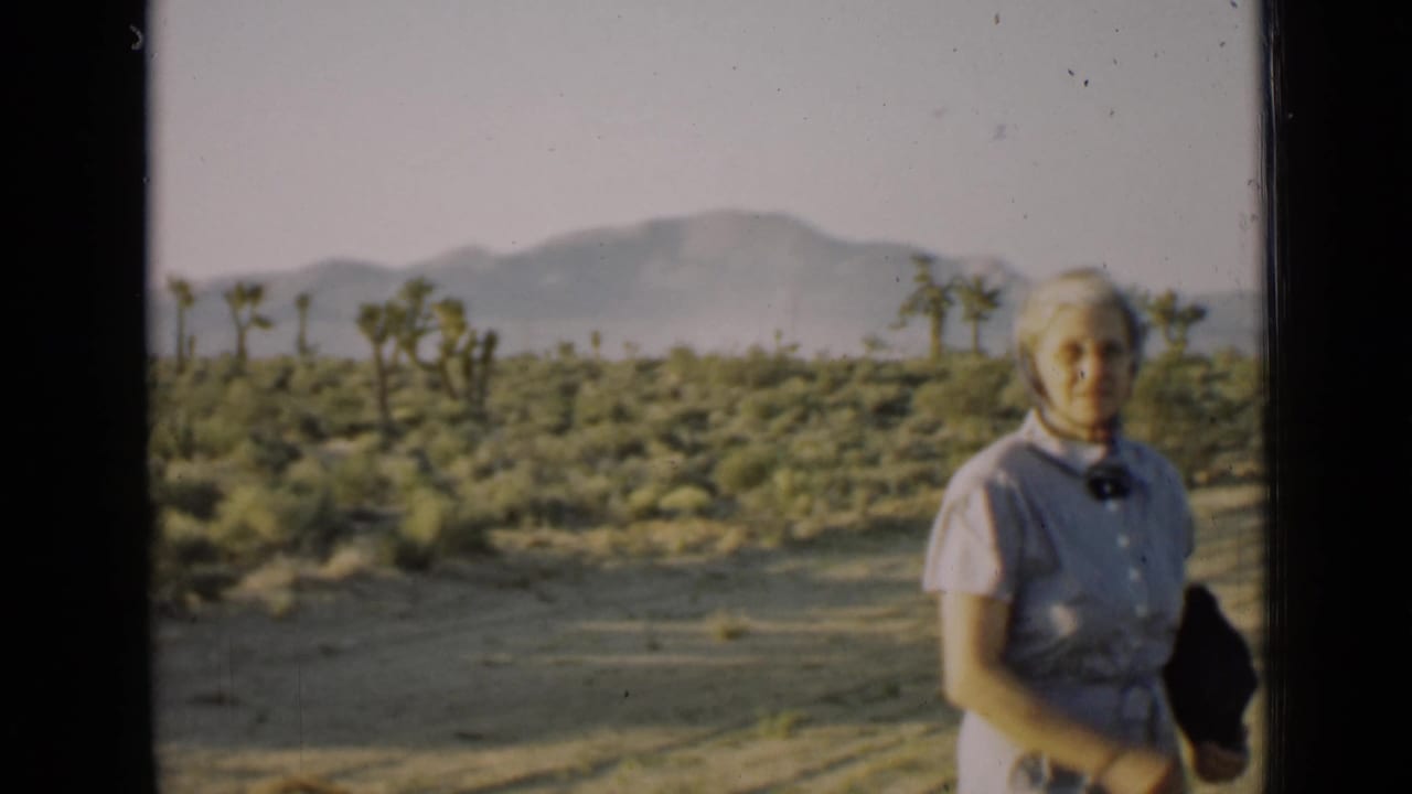 Older woman in a pale dress and headscarf standing in a desert landscape.