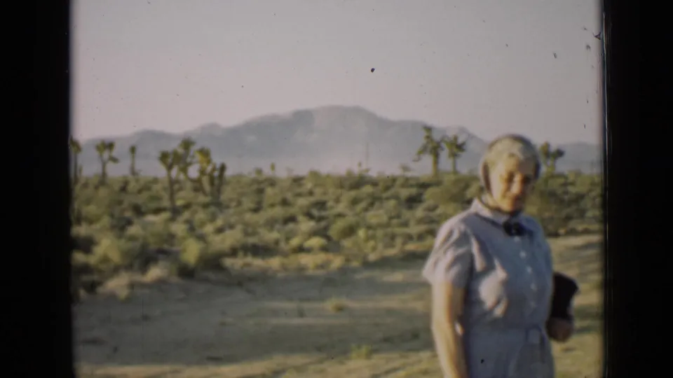 Older woman in a headscarf holds a dark hat while standing before Arizona desert plants and mountains in 1939 footage.